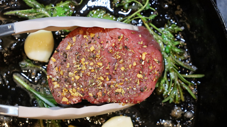 A raw filet mignon steak going into a hot pan with garlic and rosemary