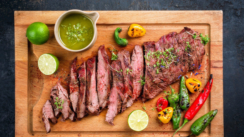 A cooked flat iron steak sliced with roasted peppers, green sauce, and limes.