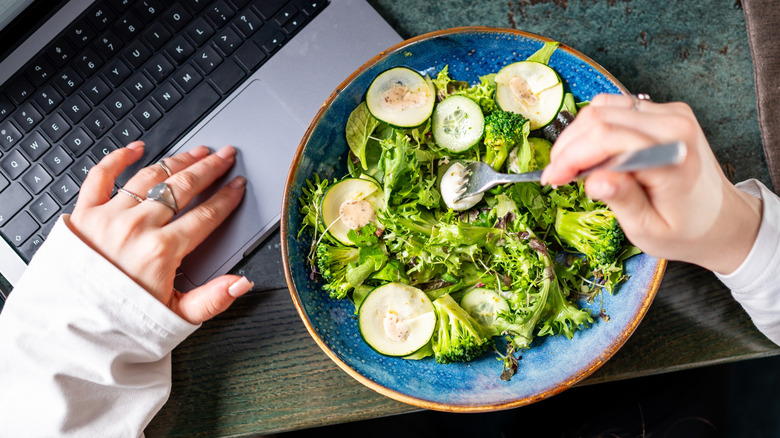 Someone sticking a fork into a bowl of green salad while also working on their computer.