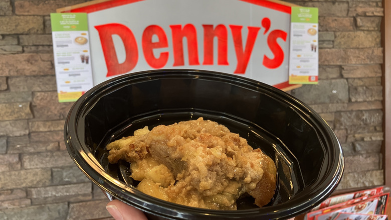 A scoop of apple pie cobbler in a black takeout bowl held in front of a Denny's Restaurant sign