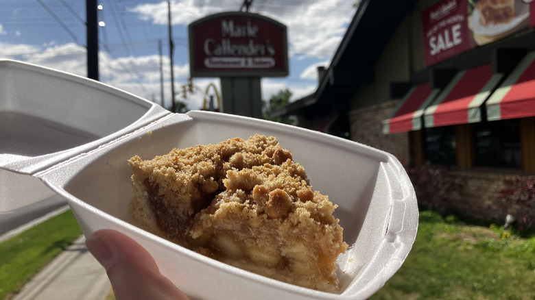 A slice of apple pie with streusel topping in a takeout carton held in front of a Marie Callender's Restaurant sign