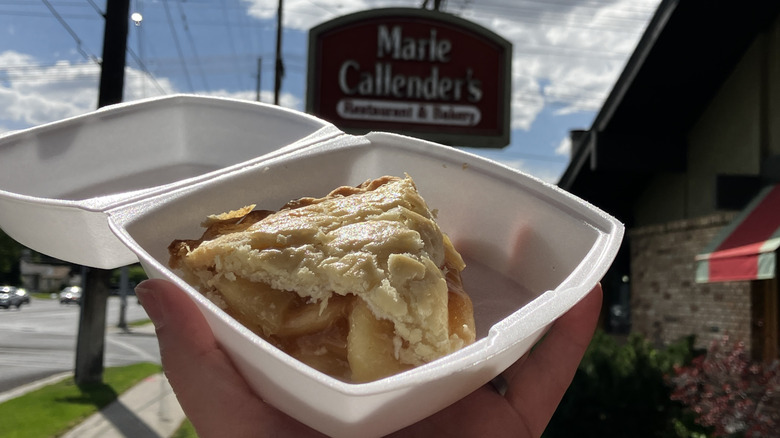 A piece of apple pie in a takeout carton held in front of a Marie Callender's restaurant sign