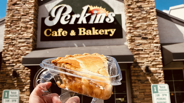 A slice of apple pie in a triangular clear plastic takeout container held in front of a Perkins Cafe & Bakery sign