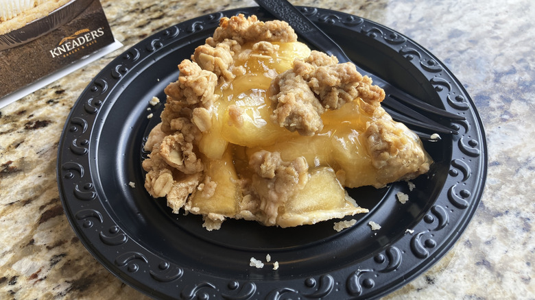 A slice of apple pie topped with oat crumble sits on a black plate on a marble countertop with a Kneaders Bakery sign in the background