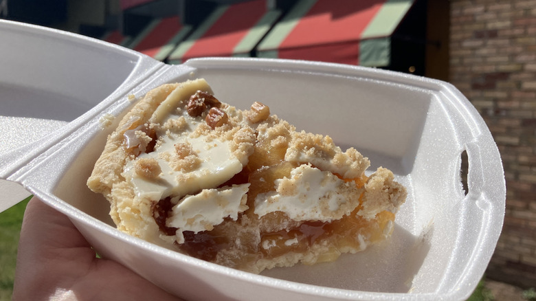 A slice of sour cream apple pie in a takeout carton held in front of a Marie Callender's Restaurant awning