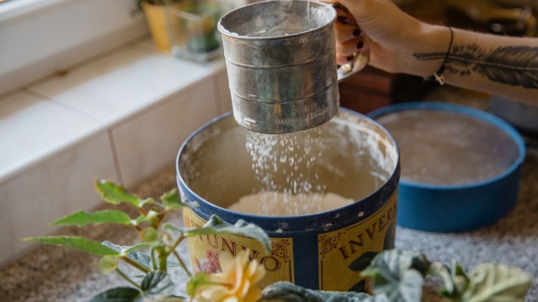 A hand holding a flour sifter over a flour canister