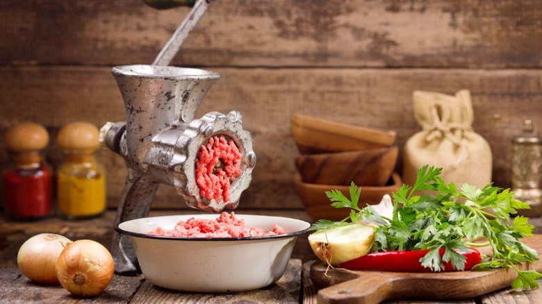 A vintage meat grinder filled with ground beef sitting on a kitchen counter with a bowl of ground beef, several yellow onions, a red chile pepper, and a pile of parsley