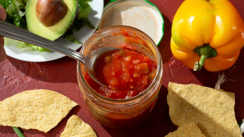 Open jar of salsa with spoon inside with tortilla chips, avocado, and a pepper nearby.