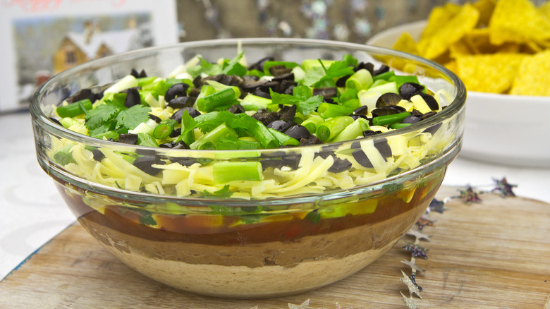 bowl of seven-layer dip on wooden surface with tortilla chips in the background
