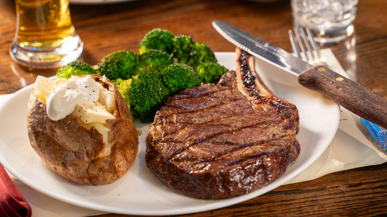 A restaurant plate with ribeye steak, broccoli, and a baked potato resting on a wooden table.