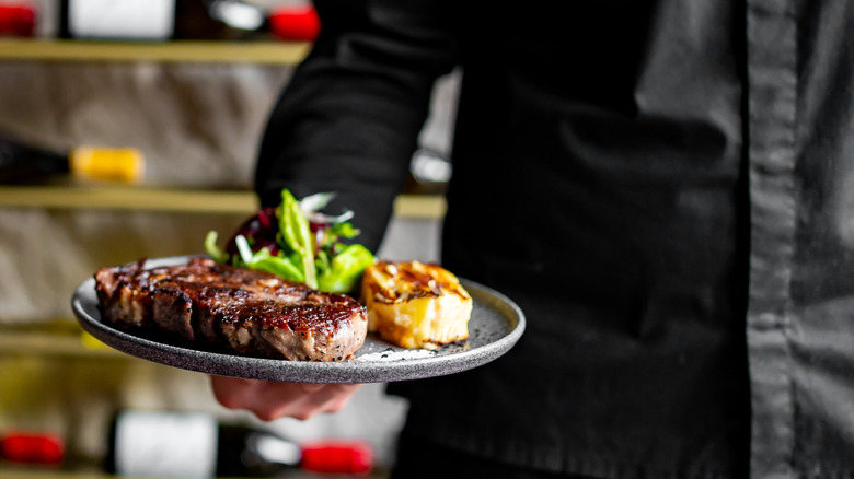 A server dressed in black holding a ribeye steak entree with scallop and salad