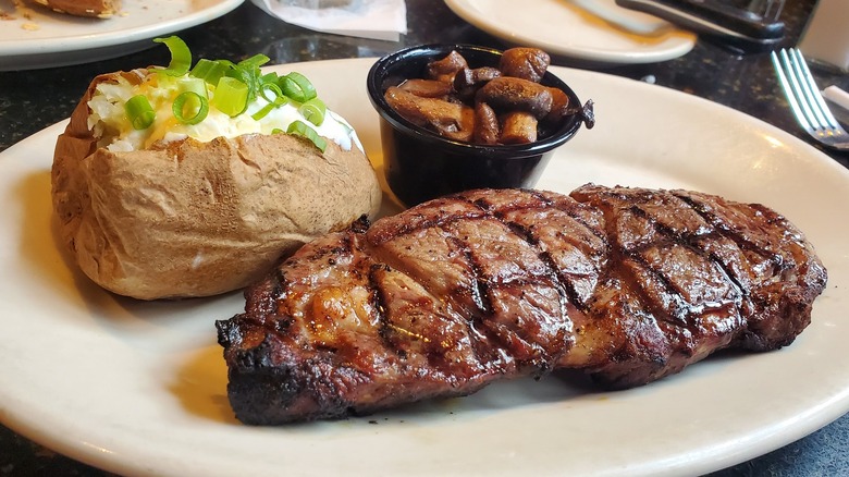 A clean white plate holding a baked potato, a ribeye steak, and a cup of mushrooms at Black Angus Steakhouse.