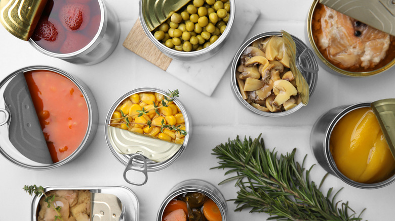 Open tin cans with different products and rosemary on white tiled table, flat lay