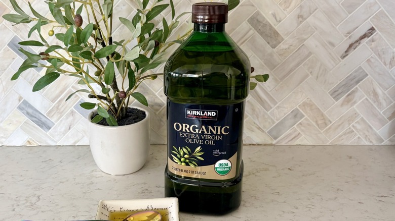 A 2-liter bottle of Kirkland Signature organic extra virgin olive oil from Costco, on counter in front of decorative olive tree in pot