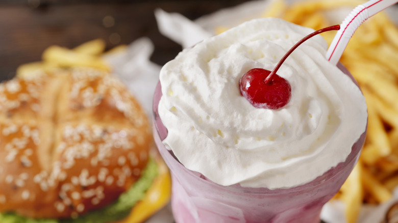 Strawberry Milkshake with whipped cream and cherrry on top, in front of a Burger and Fries