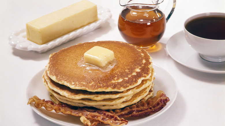 Stack Of Pancakes with butter on top and bacon on side, in front of a Stick Of Butter, Cup Of Coffee, and Pitcher Of Maple Syrup.