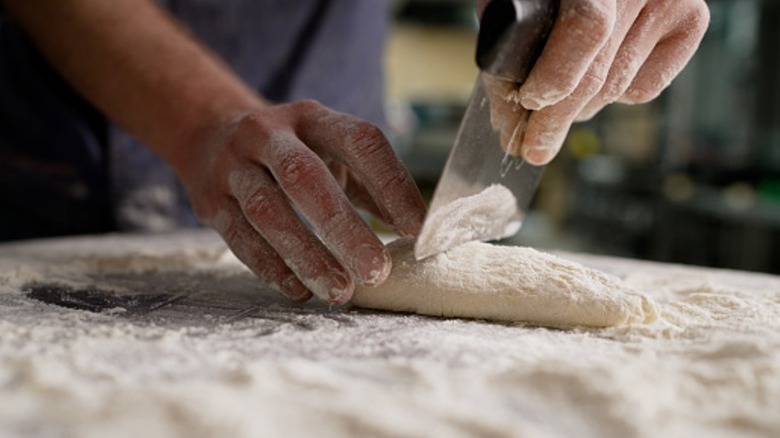 a baker uses a bench scraper to cut floured dough on floured surface