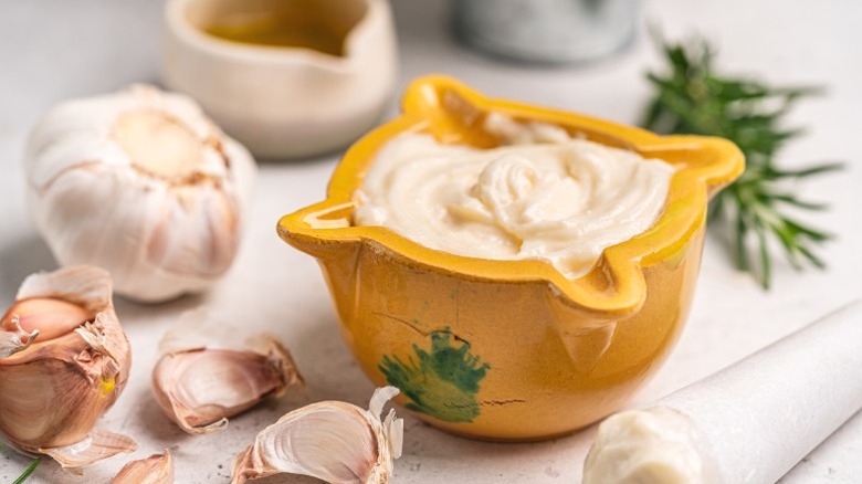 A small yellow bowl of aioli on a counter next to cloves of garlic.