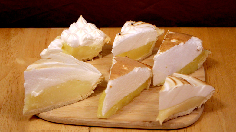 Six slices of lemon meringue pie from different brands arranged on a wooden cutting board.