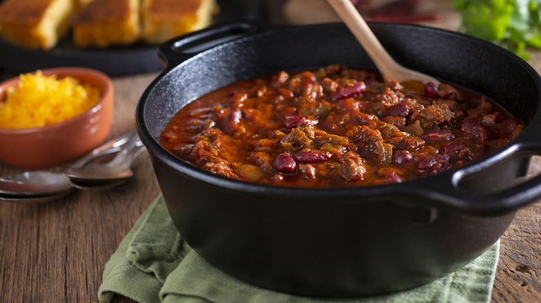 A red chili with beans in a cast iron pot on a wooden table with a bowl of cheese and spoons