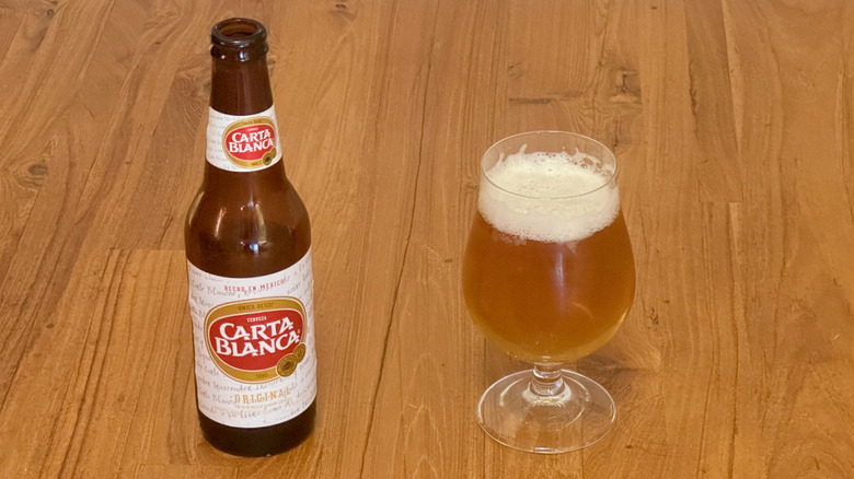 A brown bottle of Carta Blanca beer with a red and white label next to a full glass of the beer.