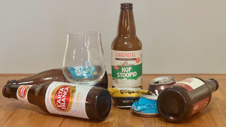 Empty beer bottles and crushed cans on a wood table around one empty bottle and beer glass, including beer brands like Carta Blanca and Lagunitas Hop Stoopid