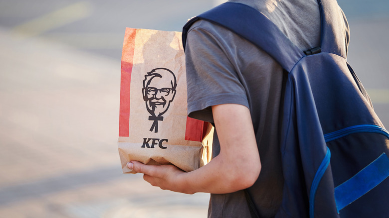 Young boy standing outdoors with book bag on while holding KFC bag