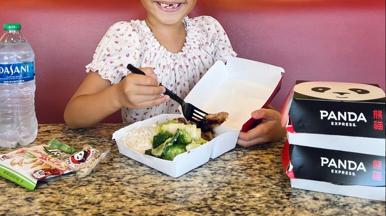 Little girl eating Panda Express' Panda Cub Meal containing broccoli and chicken with fork at table