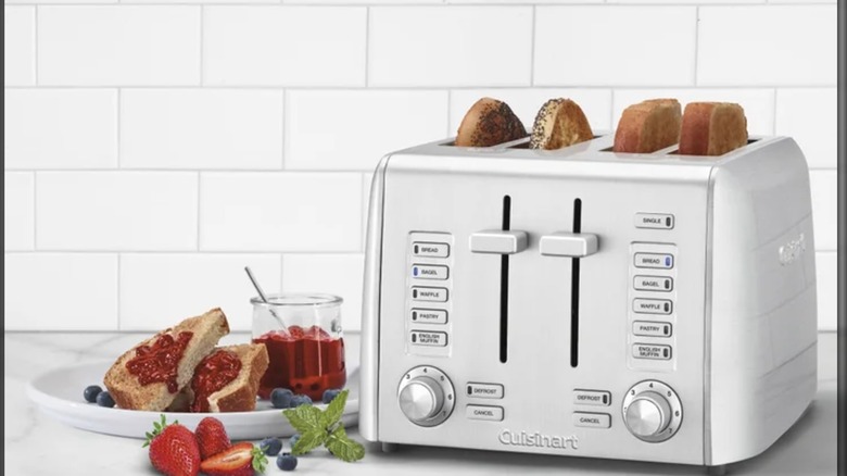 Cuisinart toaster with four slices of toast in it, next to a plate of toast and jam, and fresh fruit, on a counter in front of a tiled backsplash