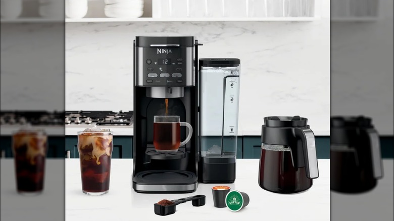 A Ninja DualBrew XL next to a glass of iced coffee, a carafe, with two pods and a scoop in front of it on a kitchen counter