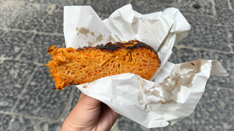 Hand holding paper wrapped frittatina with red sauce, volcanic stone Neapolitian street in background