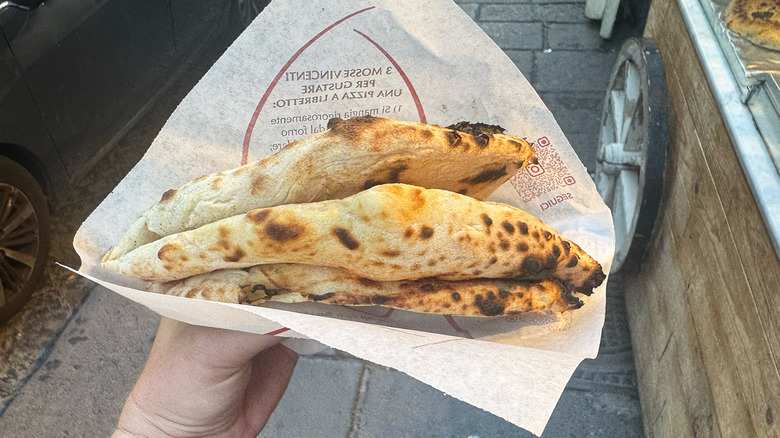 Hand holding folded pizza a portafoglio, Naples street, car, and pushcart in background