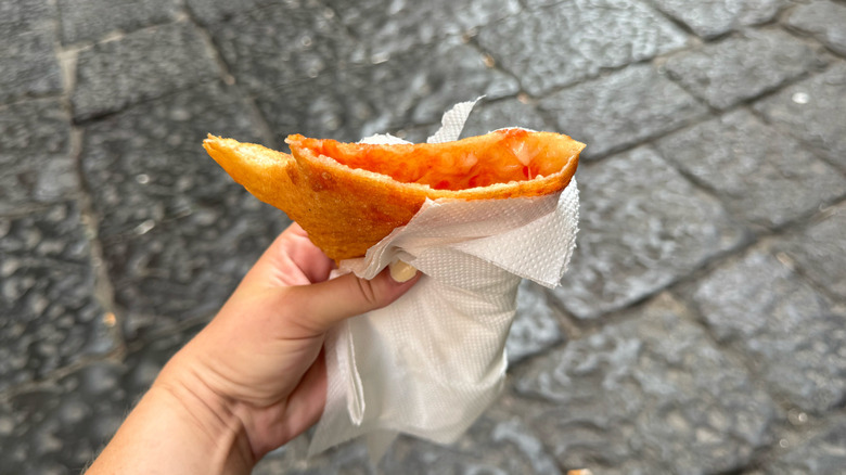 Hand holding paper wrapped pizza fritte fried pizza, volcanic stone Neapolitian street in background
