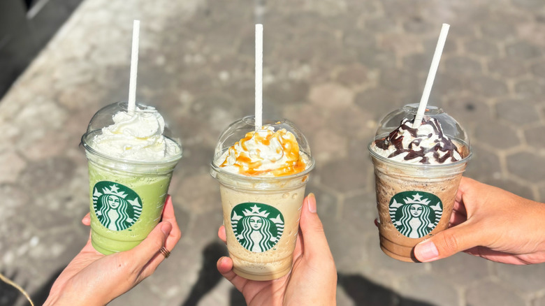 Three hands hold three different kinds of Starbucks drinks in a sidewalk-shot picture.