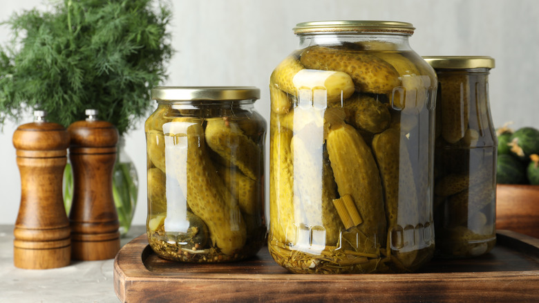 Glass jars of pickles on wooden board with pepper grinders and bunch of fresh dill in the background.