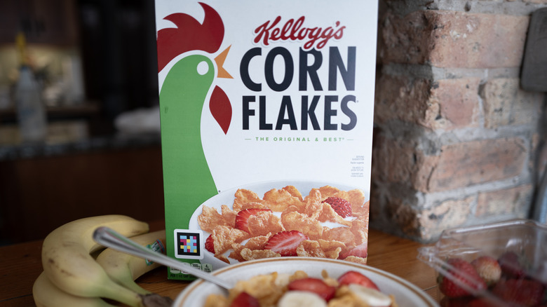 A box of Kellogg's Corn Flakes on a kitchen counter with bananas, strawberries, and bowl of cereal in the foreground