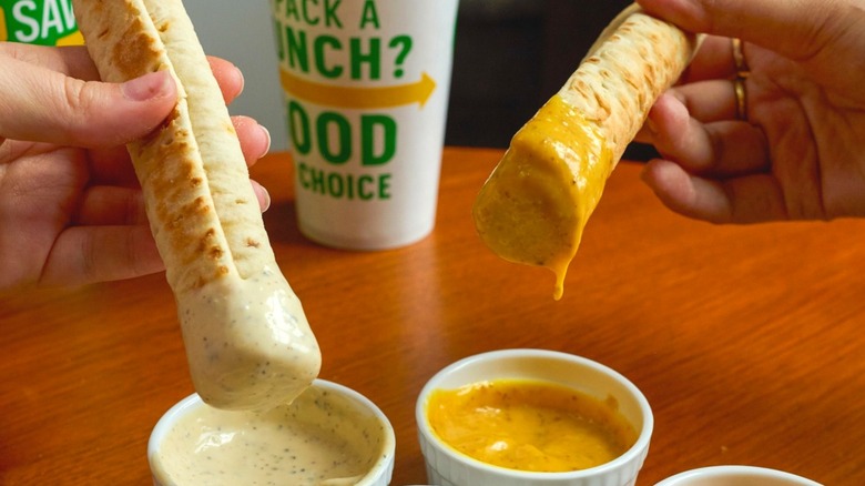 Two people hold Subway Footlong Dippers dipped into sauces