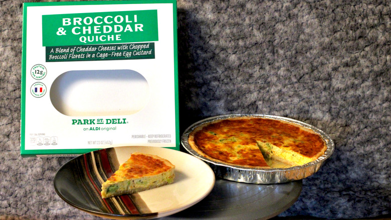 A Park Street Deli Broccoli and Cheddar Quiche in aluminum foil tray with a slice cut out and placed on a plate next to packaging