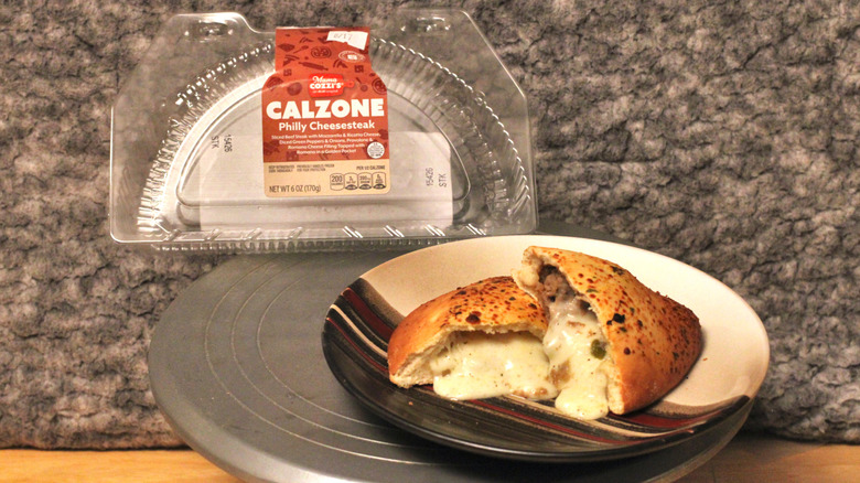 A Mama Cozzi's Philly Cheesesteak Calzone on a plate beside its plastic container