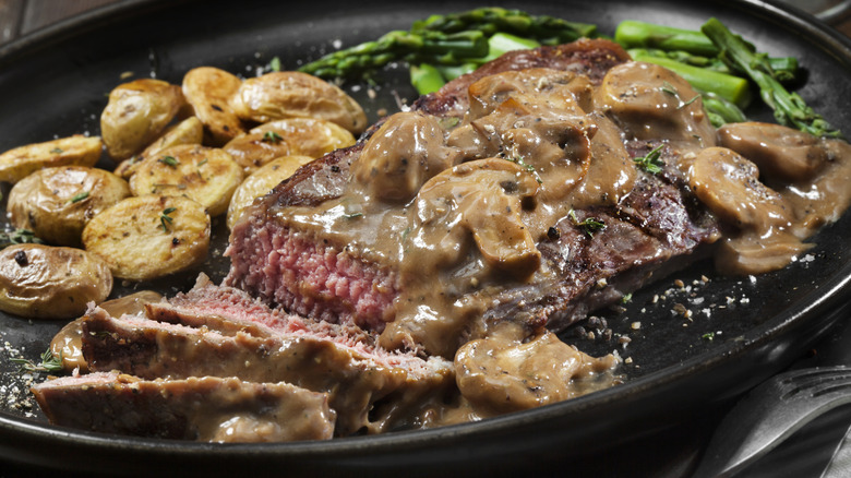 Grilled steak smothered in a creamy mushroom sauce on a black plate with potatoes and asparagus