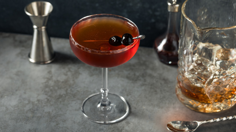 A Manhattan cocktail on a stone counter next to a jigger and a mixing glass