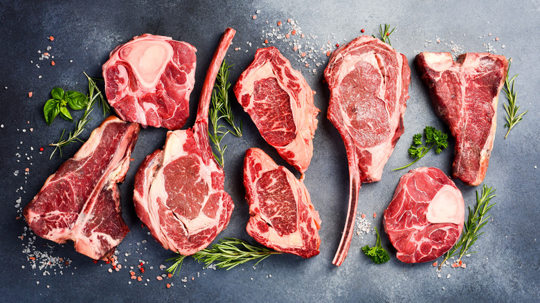 A variety of prime raw steaks on a black board surrounded by fresh herbs.