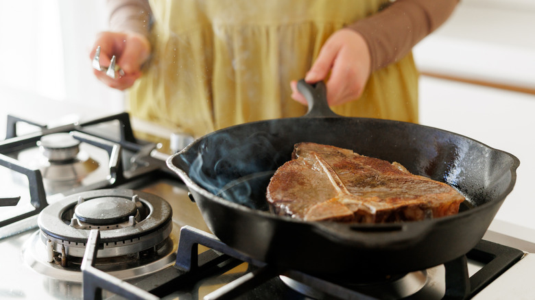 Woman searing a T-bone steak in a cast iron skillet on a gas stove, with steam rising, perfect for cooking, grilling, and gourmet food themes.