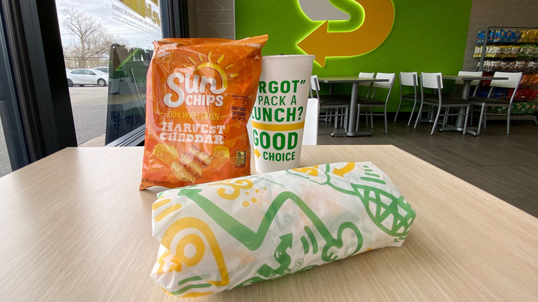 A Subway sandwich on a table with chips and a drink
