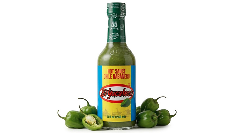 Bottle of El Yucateco Chile Habanero Hot Sauce alongside fresh green habanero peppers on white background