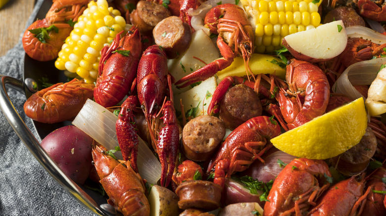 A seafood boil in a metal bowl with crawfish, corn on the cob, sausage, potatoes, and lemon wedges