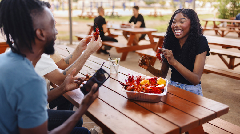 Three people at a picnic table outside sharing crawfish from a seafood boil tin on the table.