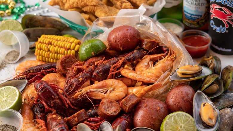 A bag of seafood boil with crawfish on a table with potatoes, corn, and limes beside bottles of beer and fried fish.