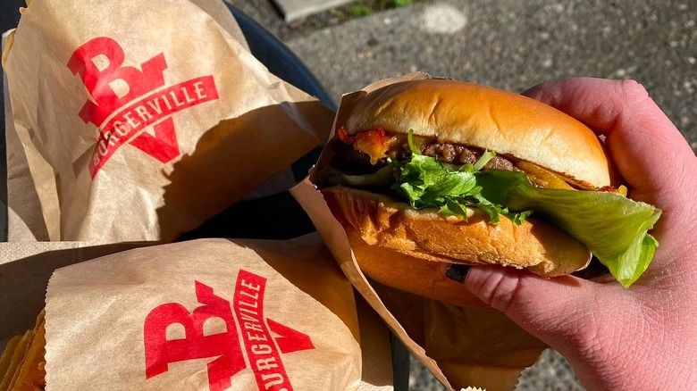 Hand holding Burgerville burger alongside small paper bags of fries