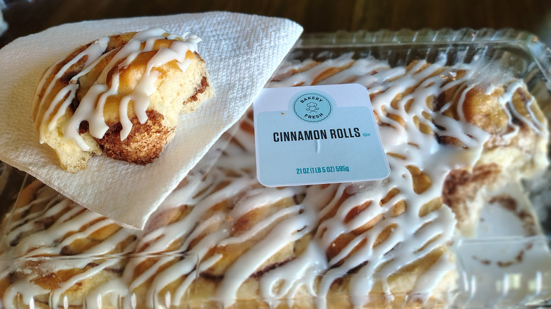 A piece of Bakery Fresh cinnamon roll with white drizzle sits on a white napkin atop an eight-pack of the pastries.